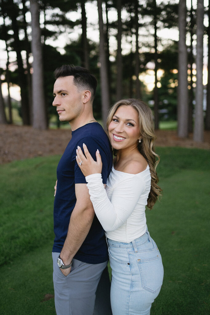 A woman in a white top and jeans smiles while standing behind a man in a navy t-shirt and gray pants. She rests her hand on his shoulder as they pose outdoors among tall trees and green grass.