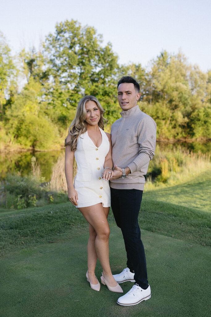 A young couple stands on green grass outdoors, with trees and a pond in the background. The woman wears a white dress and heels, and the man wears a beige sweater, dark pants, and white shoes. They smile and pose closely together.