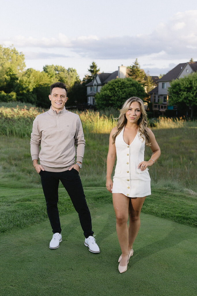 A man in a beige sweater and black pants stands next to a woman in a sleeveless white dress on a grassy lawn, with trees and houses in the background under a partly cloudy sky.