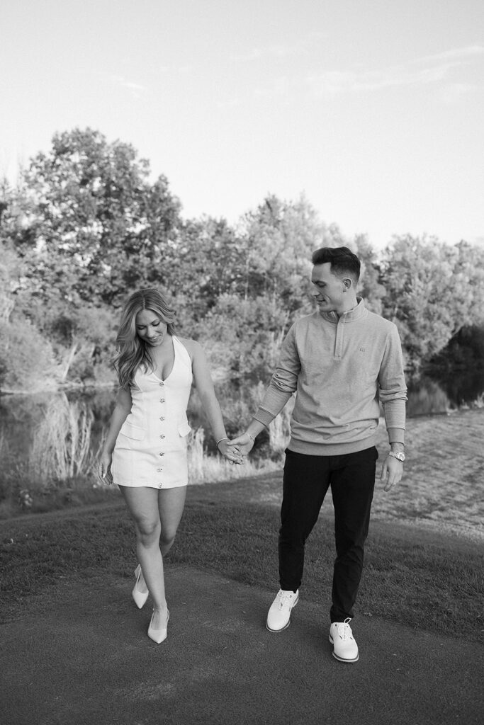 A young couple walks hand in hand outdoors near a pond, smiling at each other. The woman wears a short, sleeveless dress and heels; the man wears a light sweater, dark pants, and sneakers. Trees and grass are in the background.