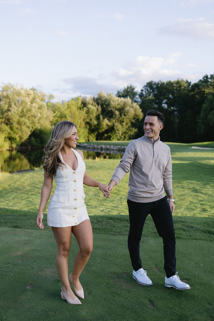 A smiling couple holds hands while walking on a grassy golf course, with trees and a pond in the background. The woman wears a white dress and heels; the man wears a light sweater, dark pants, and white sneakers.
