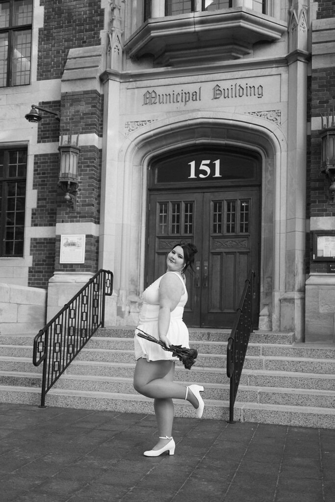 A woman in a white dress poses playfully with one leg raised, holding a bouquet in front of a building labeled Municipal Building. The scene is in black and white.
