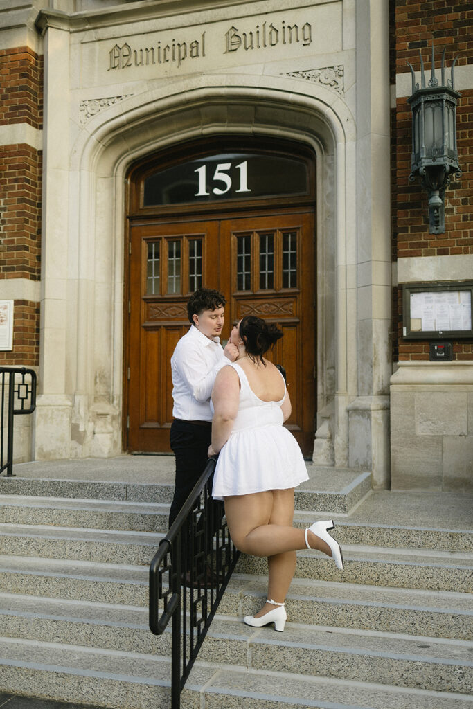 A couple stands on steps outside a building labeled Municipal Building, with one person in a white dress and heels playfully lifting a foot, facing their partner, who wears a white shirt and black pants.