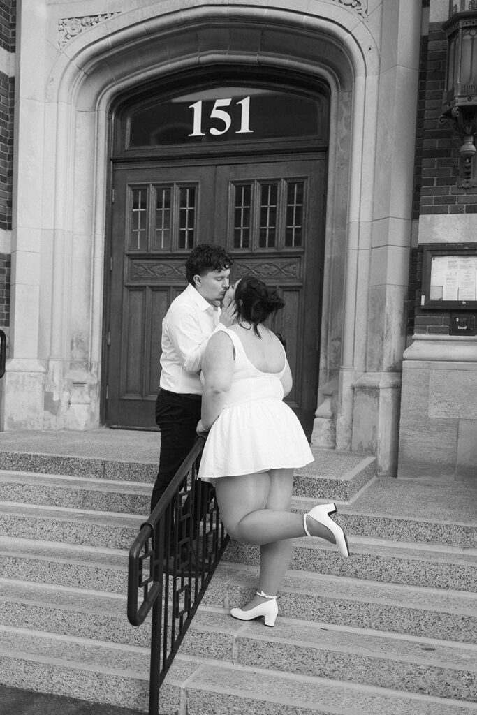 A couple dressed in white stands on stone steps outside a building with the number 151 above the door. The woman has one leg bent back playfully as they gaze at each other. The scene is in black and white.