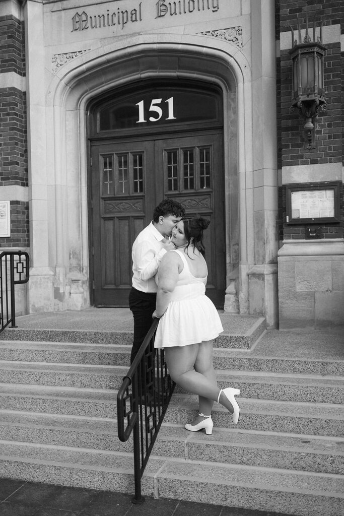 A couple dressed in white kisses on the steps of a building labeled Municipal Building above door number 151. The woman leans against a railing, and both appear happy and affectionate. The image is in black and white.