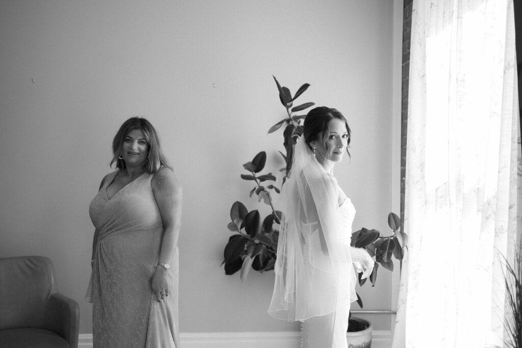 A bride in a wedding dress and veil stands near a window, looking out, while another woman in a gown stands nearby, both framed by indoor plants in a softly lit room.
