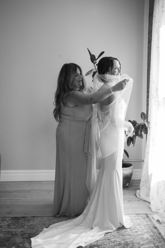 A woman in a gown helps a bride adjust her veil near a window, with soft natural light illuminating the scene. The bride stands in a long dress, and a potted plant sits in the background. The image is in black and white.