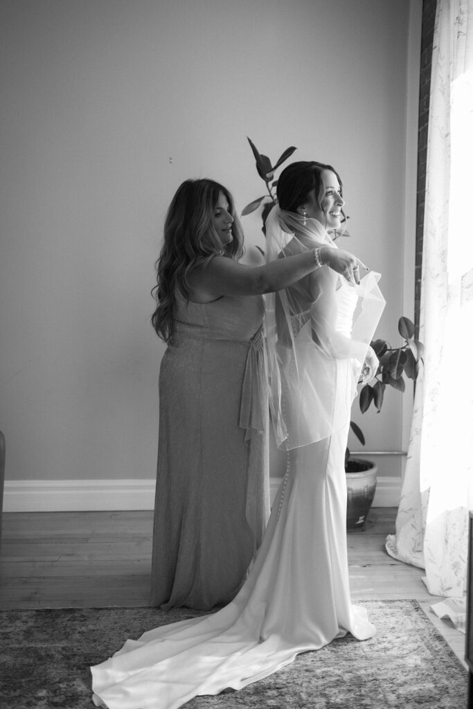 A woman in a long dress helps a bride adjust her veil as they stand near a window with sheer curtains. The bride is smiling, wearing a fitted gown with a train, while sunlight streams into the room.