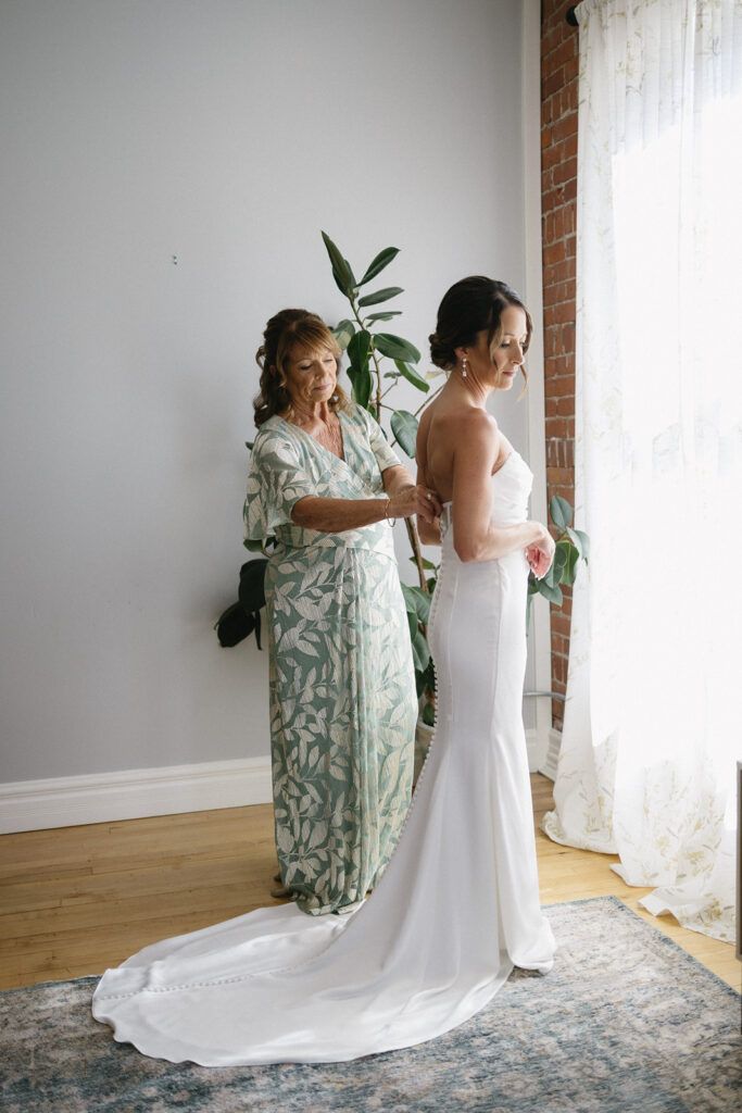 A woman in a light green floral dress helps a bride in a white gown fasten her dress near a bright window. The bride stands smiling, and the room has wooden floors, a plant, and light curtains.
