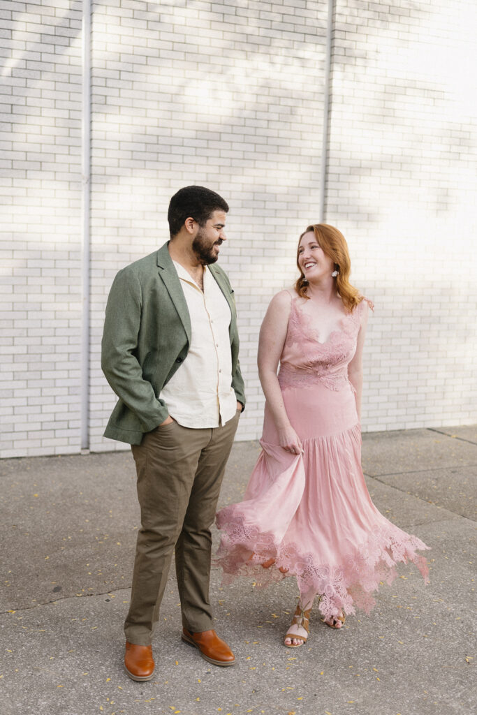 A man in a green blazer and tan pants stands next to a woman in a flowing pink lace dress. They smile at each other while walking outside in front of a white brick wall.
