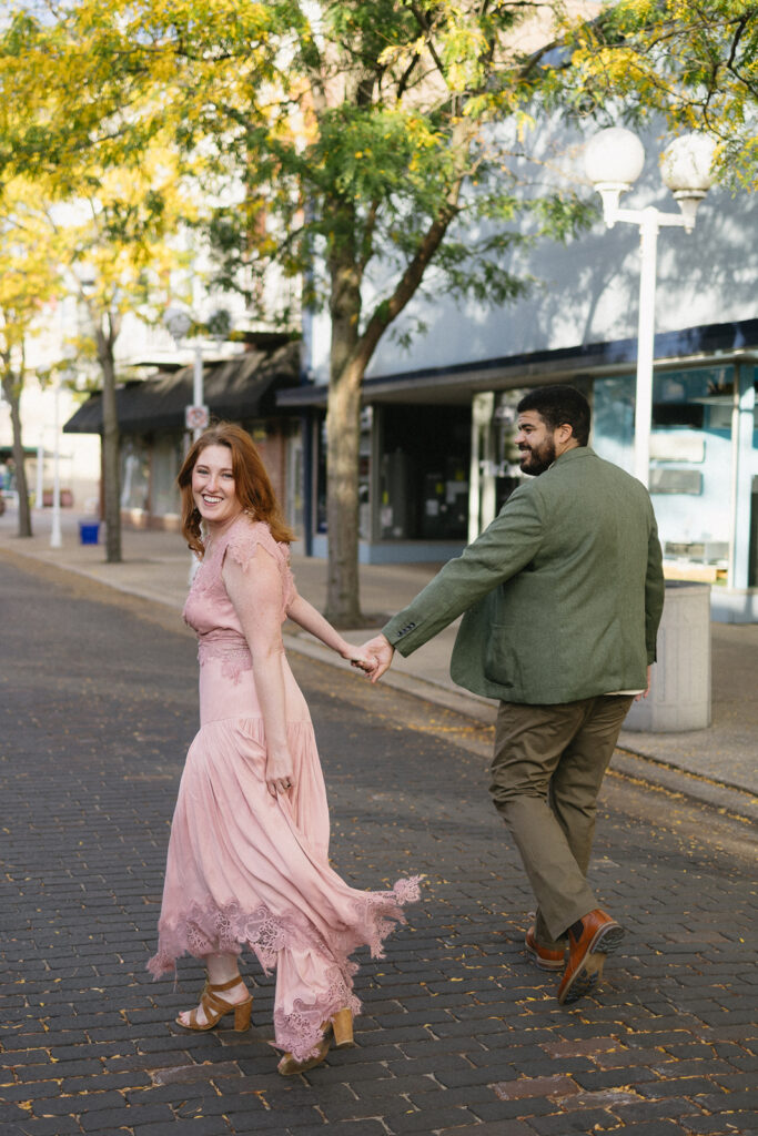 A smiling couple holds hands while walking on a tree-lined city street. The woman wears a flowing pink dress and heels, and the man wears a green jacket and brown pants. Both look back toward the camera, appearing joyful.