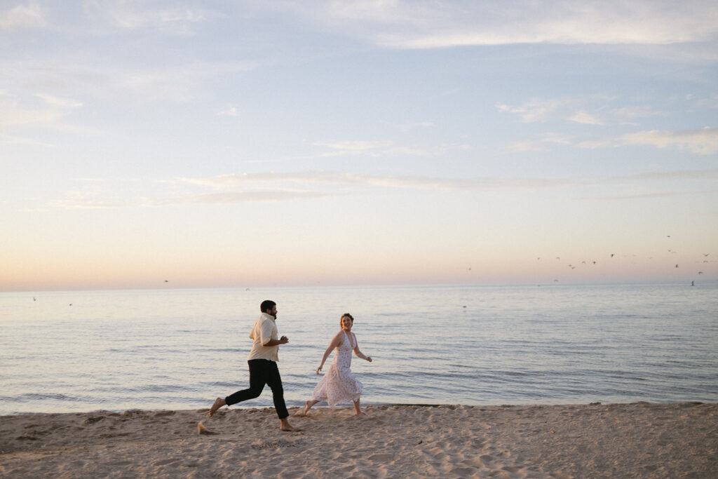 A man and woman run barefoot along a sandy beach at sunset, smiling and looking at each other, with calm water and a pastel sky in the background.