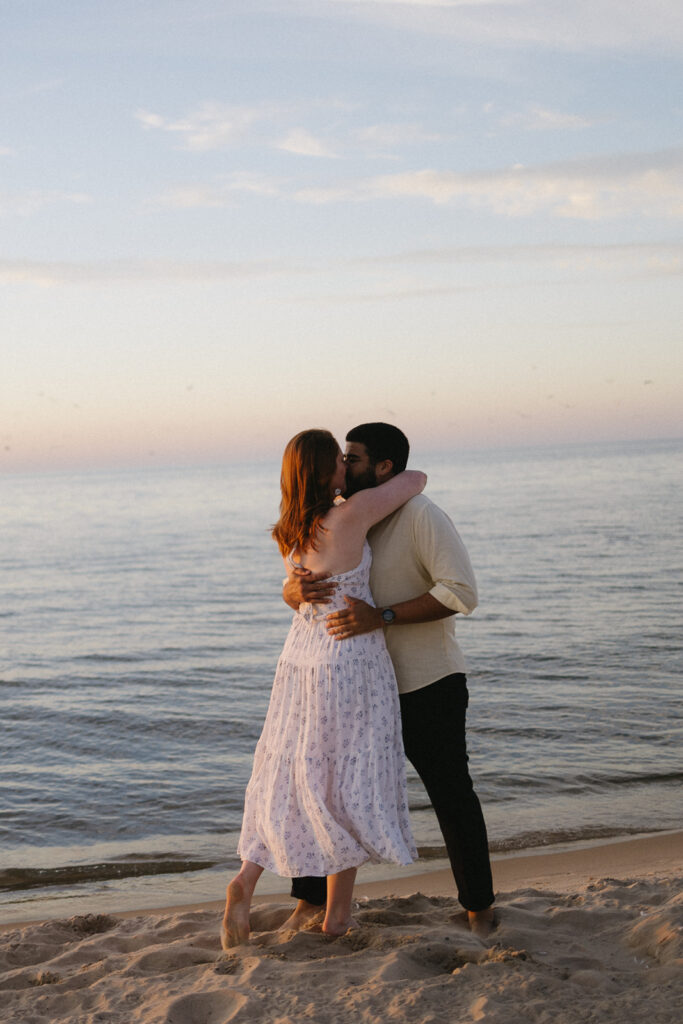 A couple embraces and kisses on a sandy beach at sunset, with gentle waves and a colorful sky in the background. The woman wears a white dress and the man wears a light shirt and dark pants.