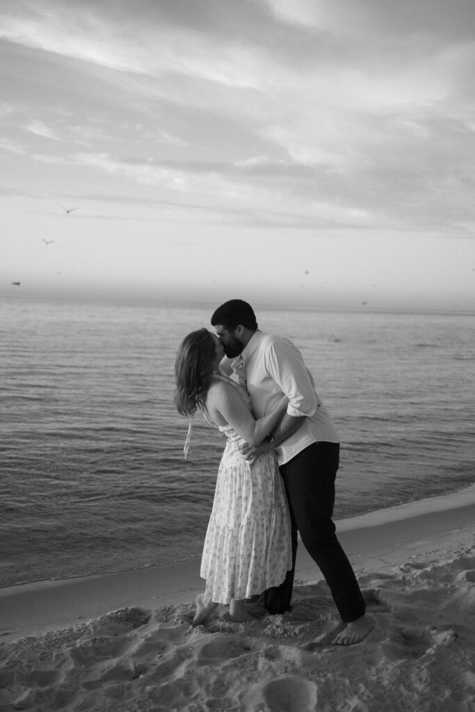 A couple stands barefoot on a sandy beach at the water’s edge, sharing a kiss. The scene is in black and white, with gentle waves and a few birds flying in the sky at sunset.