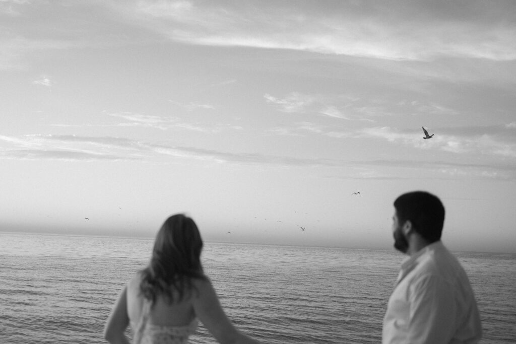 A black and white photo of a man and woman standing by the ocean, looking out at the water and sky with birds flying in the distance. The scene feels peaceful and serene.