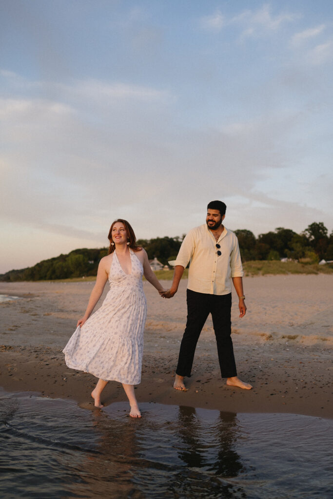 A woman in a white dress and a man in a light shirt and dark pants hold hands and walk barefoot along a sandy beach near the water, with trees and blue sky in the background.