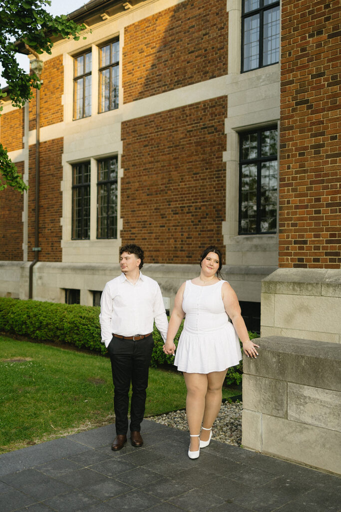 A couple stands hand-in-hand outside a brick building with large windows. Both wear white outfits; the woman wears a short dress and heels, while the man wears a shirt and pants. They stand on a stone path near green shrubs.