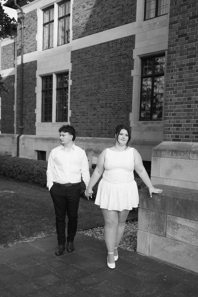 A couple holds hands while walking outside a brick building. The person on the left wears a white shirt and dark pants; the person on the right wears a sleeveless white dress and heels, resting one hand on a stone ledge. The photo is black and white.
