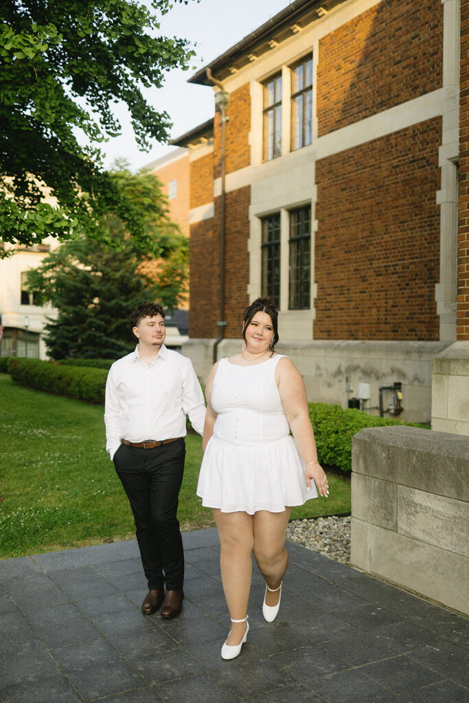A couple walks outside by a brick building. Both wear white outfits; the woman wears a sleeveless dress and heeled shoes, while the man wears a button-up shirt with dark pants and shoes. They hold hands and smile in the evening light.