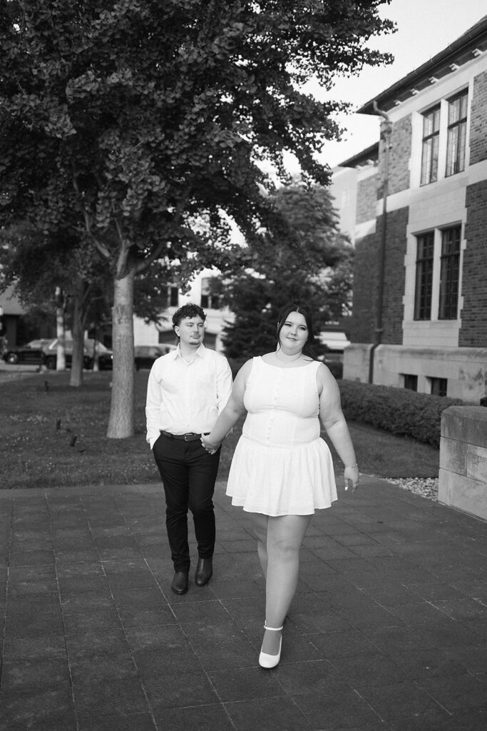A couple walks hand-in-hand on a sidewalk. The woman, in a white sleeveless dress, walks in front, looking at the camera and smiling. The man, in a white shirt and dark pants, follows slightly behind. Trees and a building line the street.
