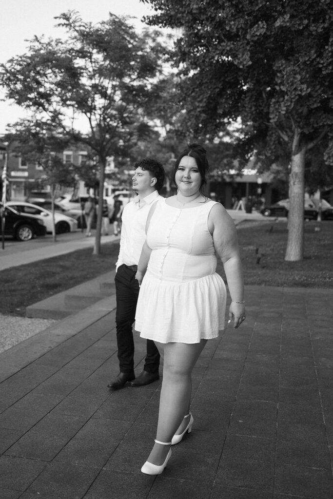 A woman in a white dress and heels walks confidently on a paved path, smiling at the camera. A man in a white shirt and dark pants walks behind her. Trees and parked cars line the street in the background. The photo is black and white.