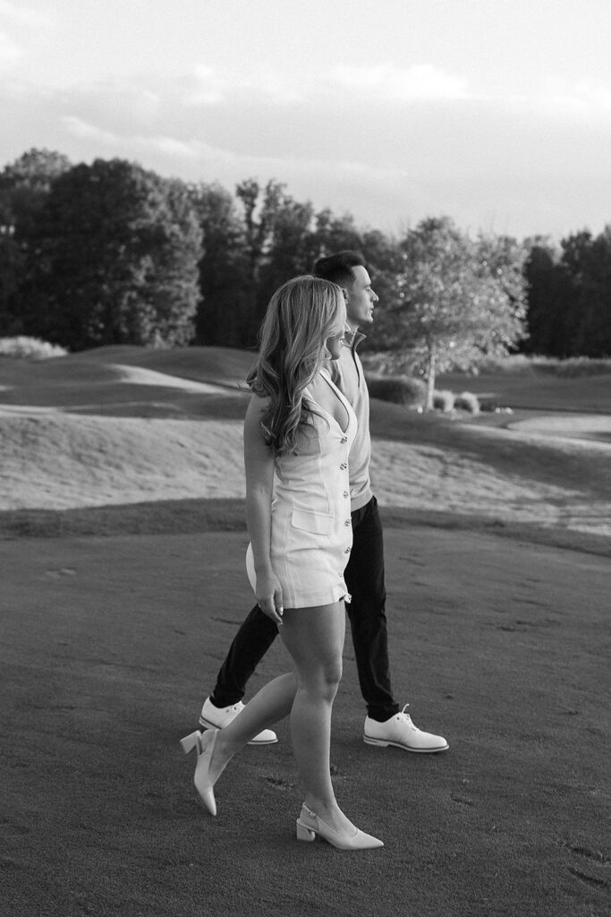 A man and woman walk side by side on a grassy area, both dressed in light-colored, semi-formal clothing. The background features trees and open landscape. The image is in black and white.