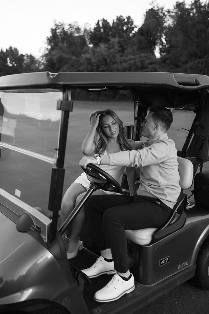 A couple sits together in a golf cart on a grassy course, smiling and enjoying each others company. The photo is in black and white, with trees visible in the background.