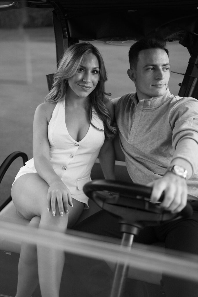 A woman in a white dress and a man in a light sweater sit together in a golf cart. The woman smiles at the camera while the man looks off to the side, holding the steering wheel. The photo is in black and white.