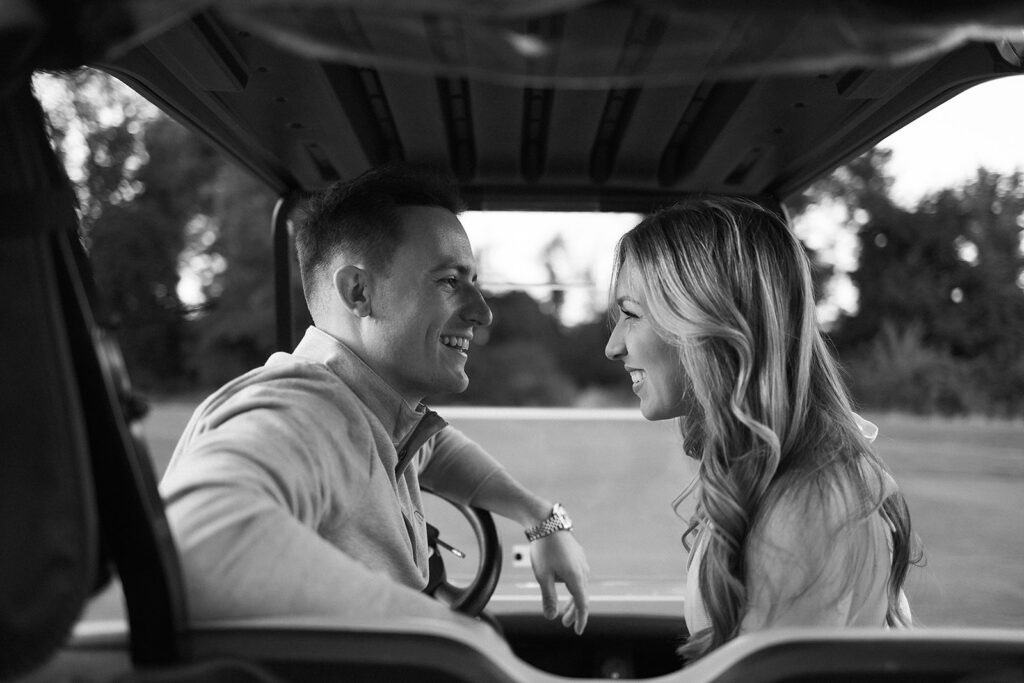 A man and woman sit facing each other in a golf cart, smiling warmly and making eye contact. The background shows trees and an open field. The image is in black and white.