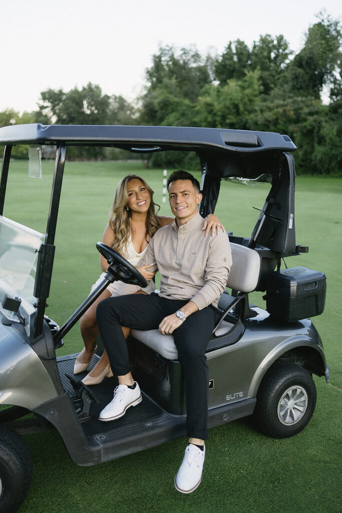 A smiling couple sits together in a golf cart on a green golf course. The woman has long hair and wears a light dress, while the man wears a tan sweater and black pants. Trees are visible in the background.