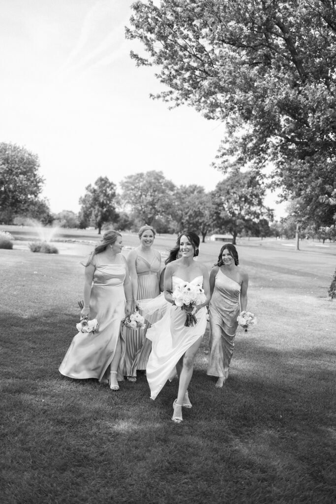Four women in elegant dresses walk across a grassy area outdoors, holding bouquets. The woman in front wears a white dress and smiles while walking barefoot. Trees and a fountain are visible in the background. The image is black and white.