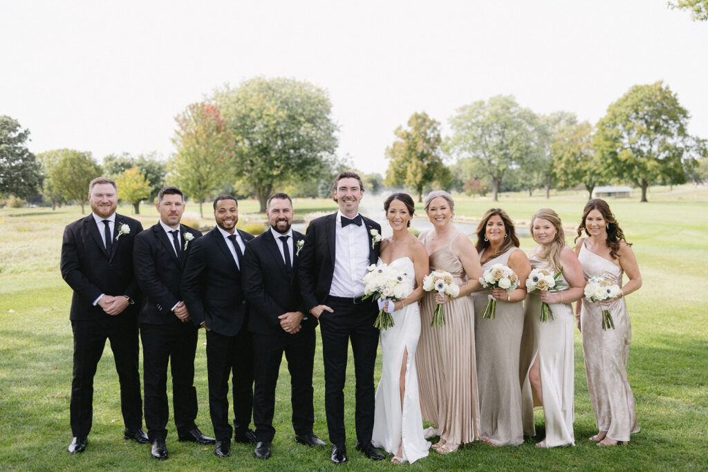 A wedding party poses outdoors on grass. Four men in black suits and ties stand beside the groom in a black tuxedo; five women in pale dresses, holding white bouquets, stand beside the bride in a white gown. Trees are in the background.