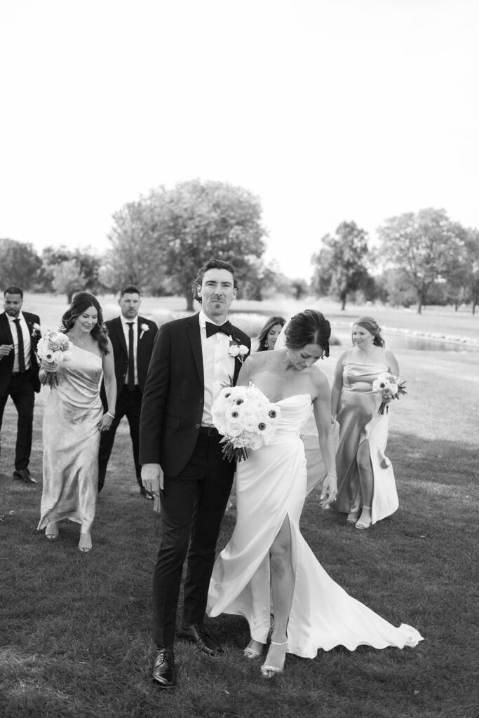 A bride and groom walk on grass with their wedding party. The groom wears a tuxedo and the bride wears a strapless gown, holding a bouquet. Bridesmaids and groomsmen, dressed formally, follow behind. The scene is outdoors in daylight.