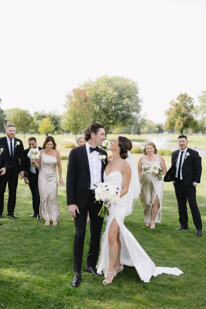 A bride and groom stand close together outdoors, smiling at each other, surrounded by their wedding party dressed in formal attire with bouquets, on a grassy lawn with trees in the background.