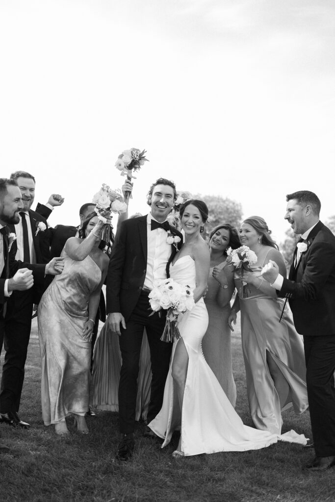 A joyful bride and groom stand together, surrounded by their smiling wedding party dressed in formal attire, raising bouquets and cheering outdoors.