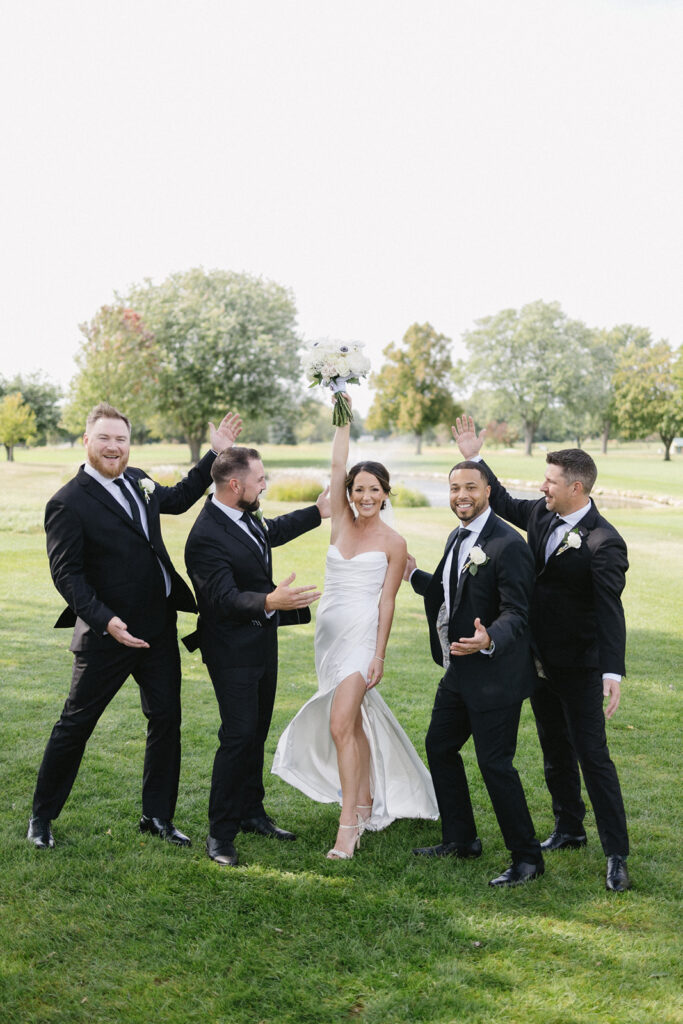 A bride in a white dress holds her bouquet high and smiles, surrounded by four groomsmen in black suits who gesture toward her joyfully, all posing outdoors on a grassy lawn.