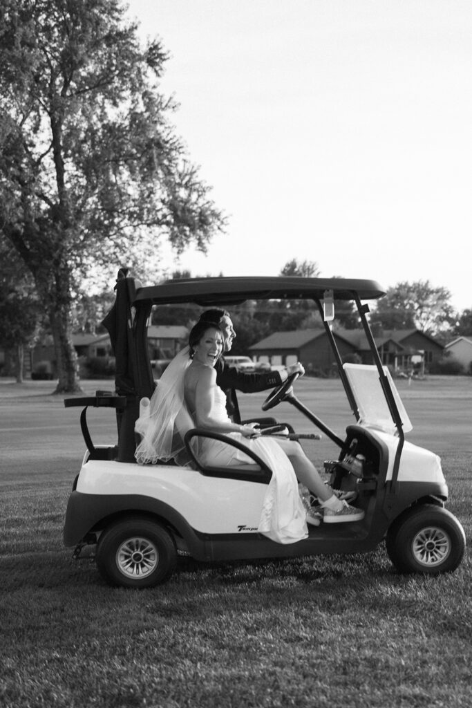 A bride in a wedding dress and veil sits smiling in the driver’s seat of a golf cart on a grassy area, with trees and houses in the background. The photo is in black and white.
