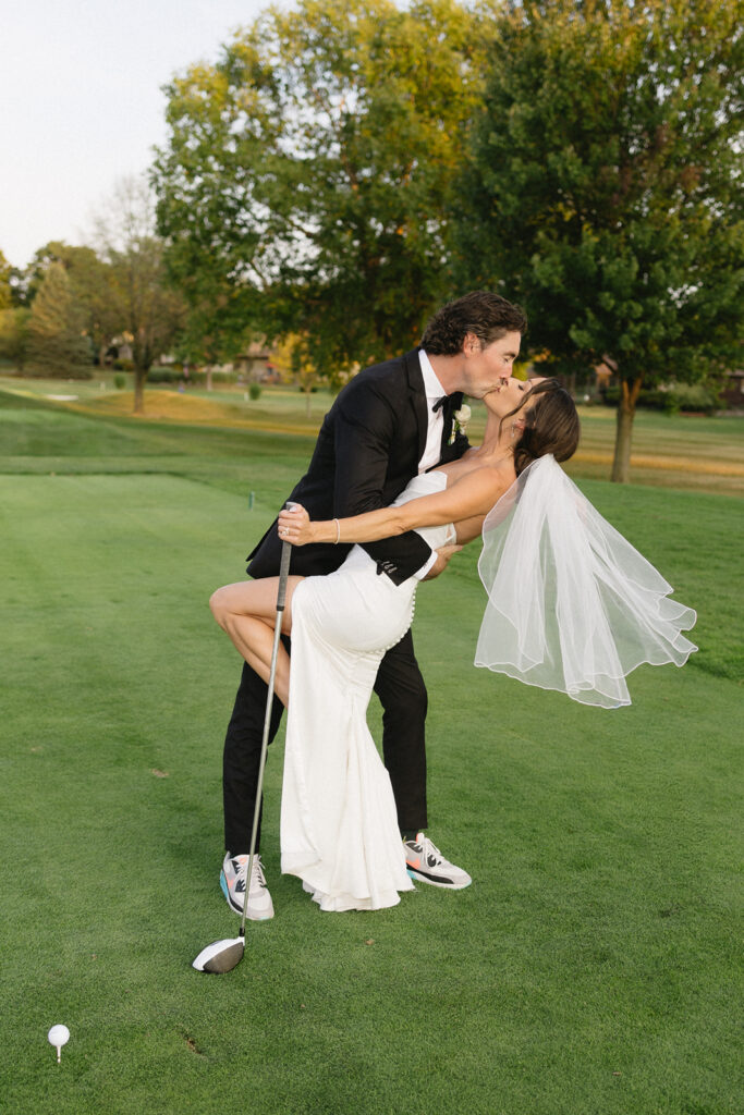 A newlywed couple shares a kiss on a golf course; the groom in a black suit dips the bride, who wears a white dress and veil, while holding a golf club. The green and trees are visible in the background.