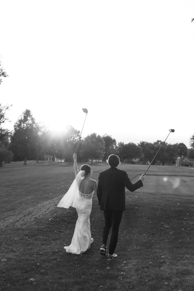 A bride and groom walk hand in hand on a golf course at sunset, both holding golf clubs raised in the air, with trees in the background.