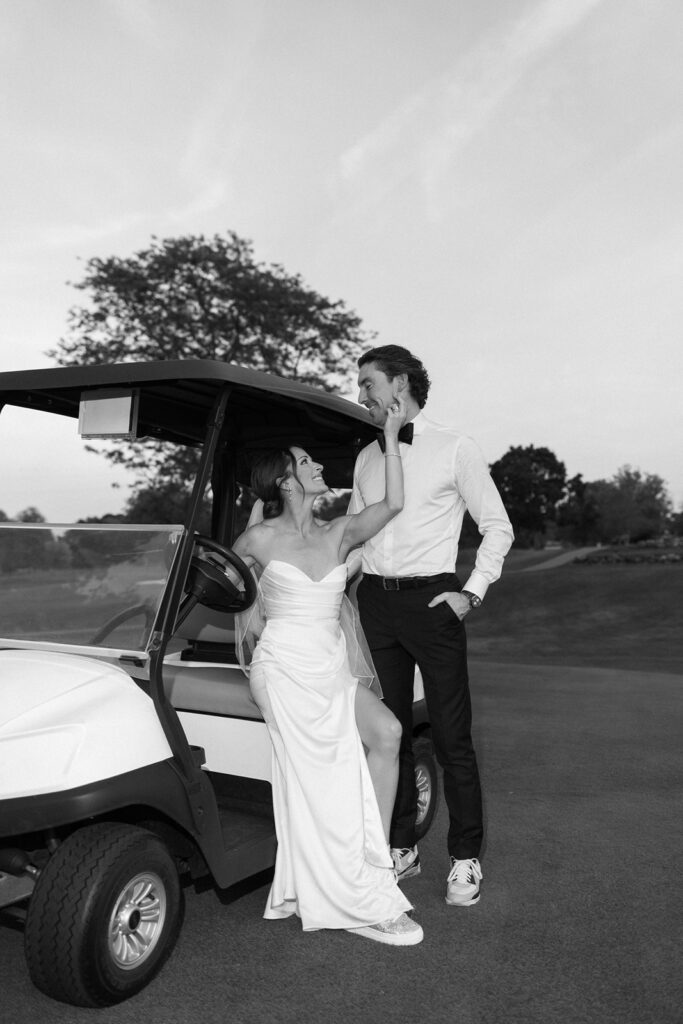 A bride in a strapless dress and veil sits on a golf cart, smiling and touching the face of a groom in a white shirt and dark pants. They look at each other affectionately outdoors on a golf course.