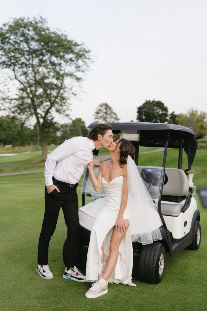 A bride in a white dress and veil sits on a golf cart, kissing a groom in a white shirt and black pants. Both wear sneakers. They are outdoors on a golf course with green grass and trees in the background.