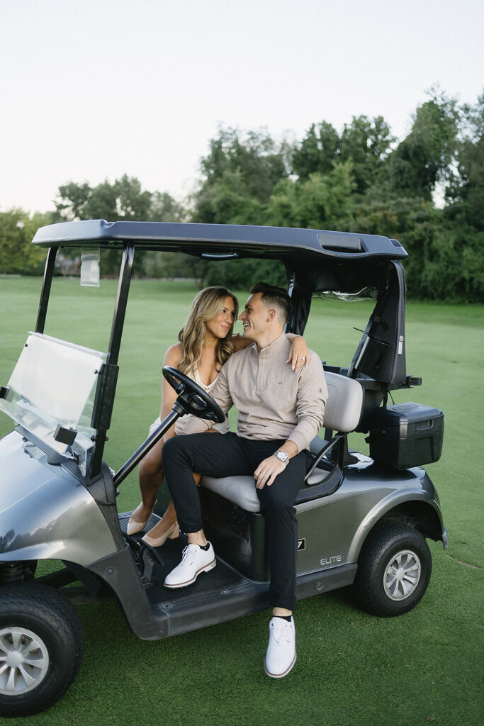 A woman and man sit closely together on a golf cart on a green golf course, smiling at each other with trees in the background.