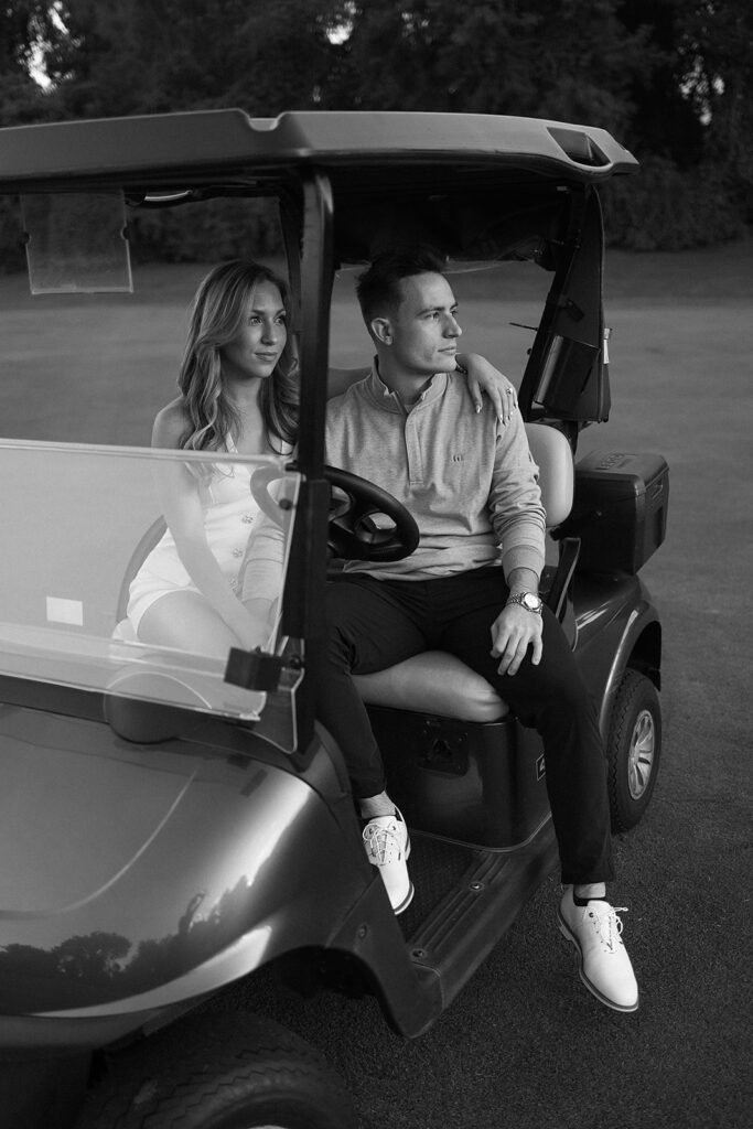 A woman and a man sit together in a golf cart on a grassy course. The woman has her arm around the man’s shoulders. Both look thoughtfully into the distance. The image is in black and white.