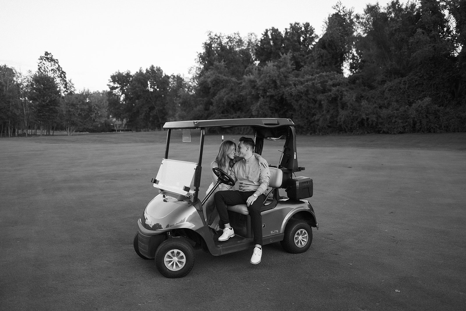 A couple sits closely together in a golf cart on an open golf course, sharing a kiss. Trees border the background, and the scene is in black and white.