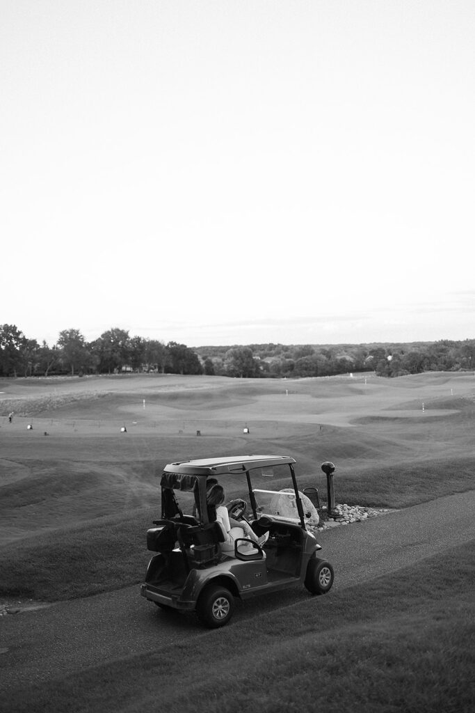 A person drives a golf cart along a paved path on a golf course, with golf clubs in the back and expansive grassy fairways in the background under a clear sky. The image is in black and white.
