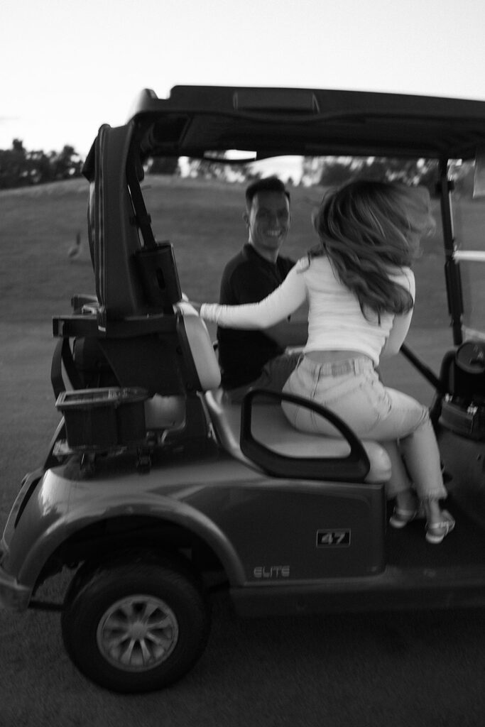 A black and white photo of a man driving a golf cart while a woman with long hair sits on the back, turning her head. Both appear to be enjoying themselves on a grassy course.