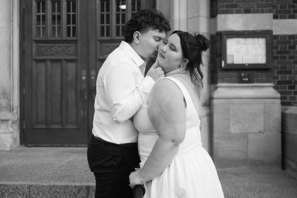 A couple dressed in white shares an intimate moment outside a building; the man gently kisses the womans temple as she smiles softly, both appearing happy and affectionate. The scene is in black and white.