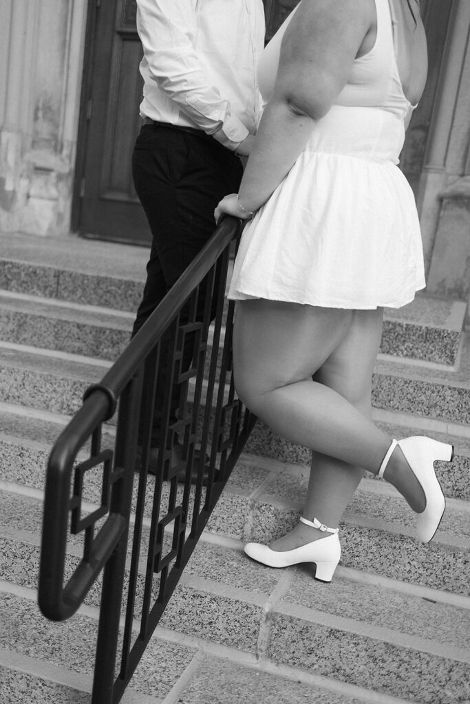 A couple stands close together on stone steps. The woman, wearing a white dress and heels, leans on a black railing, while the man, dressed in a white shirt and dark pants, stands facing her. The image is in black and white.