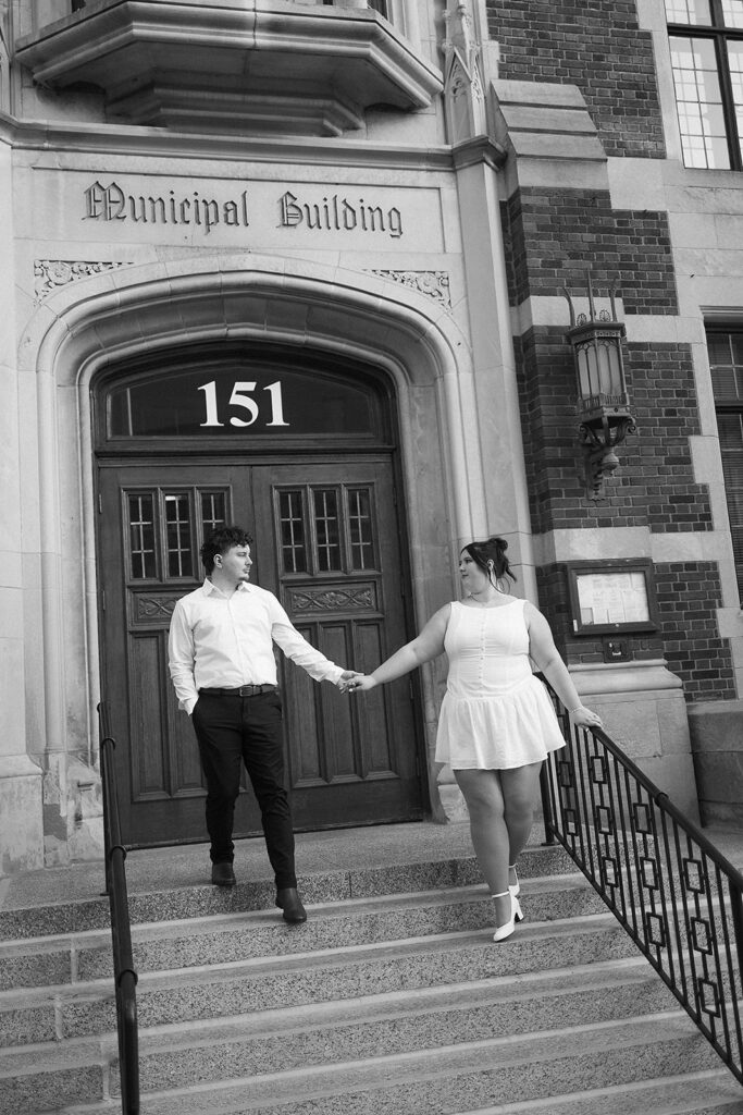 A couple holds hands on the steps of a historic building labeled Municipal Building 151. They look at each other, both smiling, dressed in light-colored clothing. The photo is black and white.