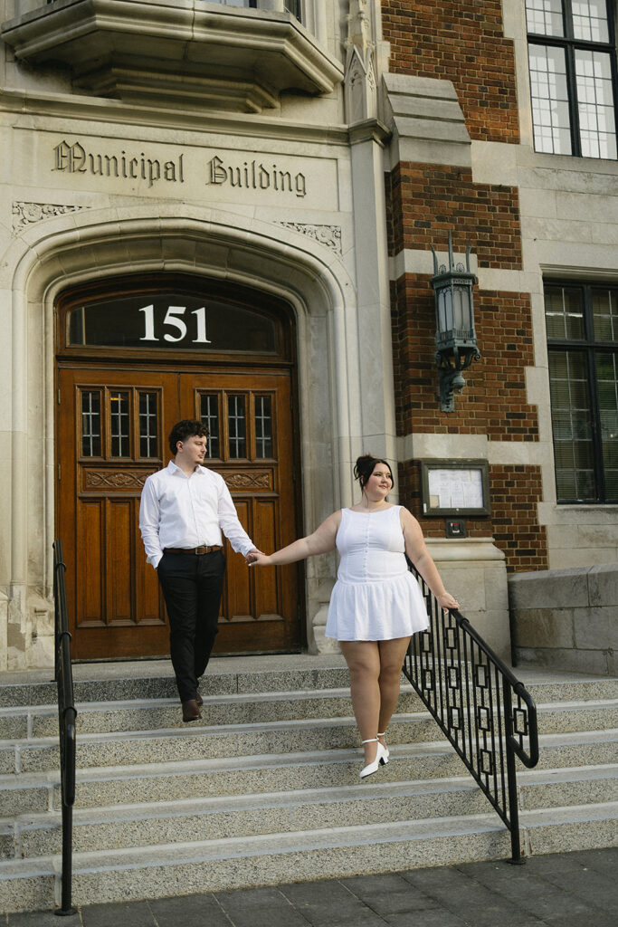 A couple holds hands and smiles while standing on the steps of a municipal building, both dressed in white outfits. The building has Municipal Building above the door and the number 151 displayed.